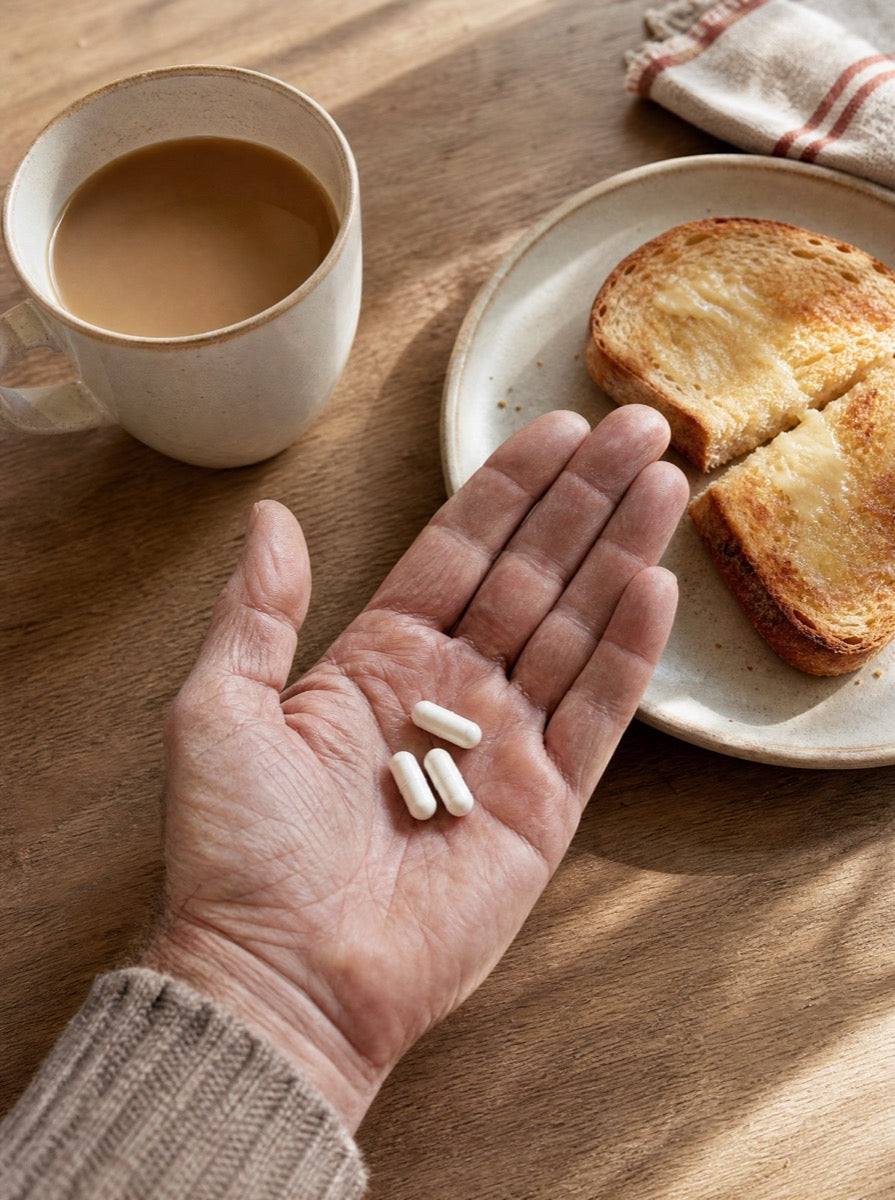 Three capsules in an open palm next to coffee and toast